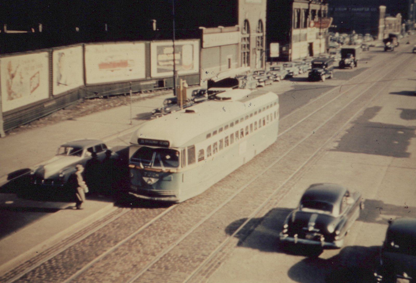 Elevated view of a green and cream streetcar on a cobblestone street with double tracks. People stand at its front beside a parked car, dark sedans pass in the next lane, and billboards and old buildings line the far side.