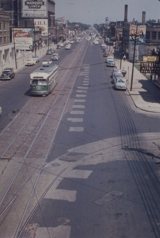 A vintage green and cream Chicago streetcar with number 4059 travels down a wide, sunlit street lined with 1950s cars and storefronts.