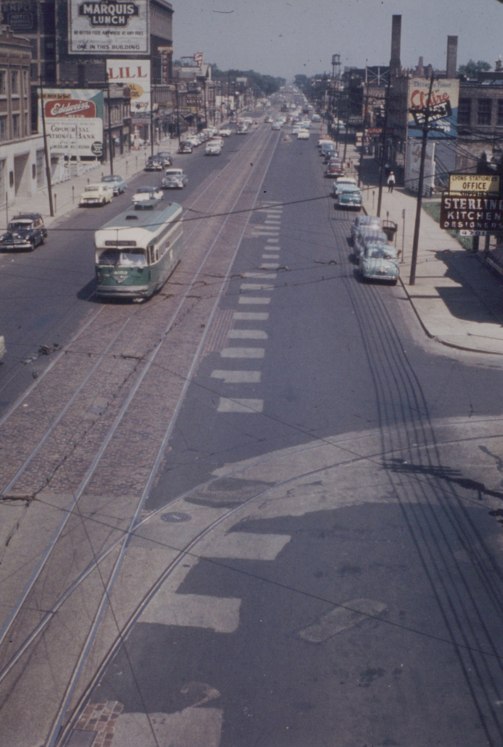 A vintage green and cream Chicago streetcar with number 4059 travels down a wide, sunlit street lined with 1950s cars and storefronts.