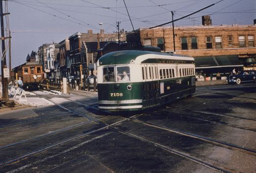 A green and cream streetcar numbered 7156 crosses a street intersection with diagonal track crossings, while an old wooden elevated train car stands on ground-level tracks behind barricades at left under overhead wires.