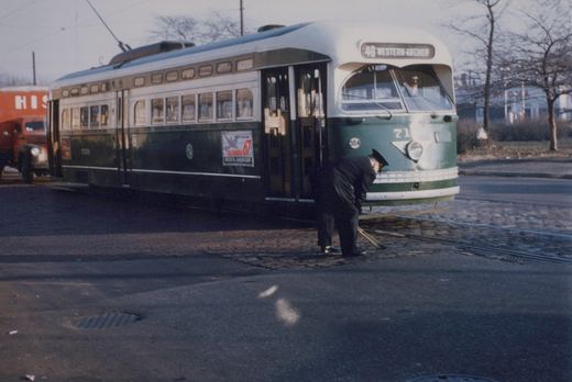 A green and cream streetcar signed 49 Western-Archer stands on cobblestone track while a uniformed crew member bends down to throw a switch with an iron bar.