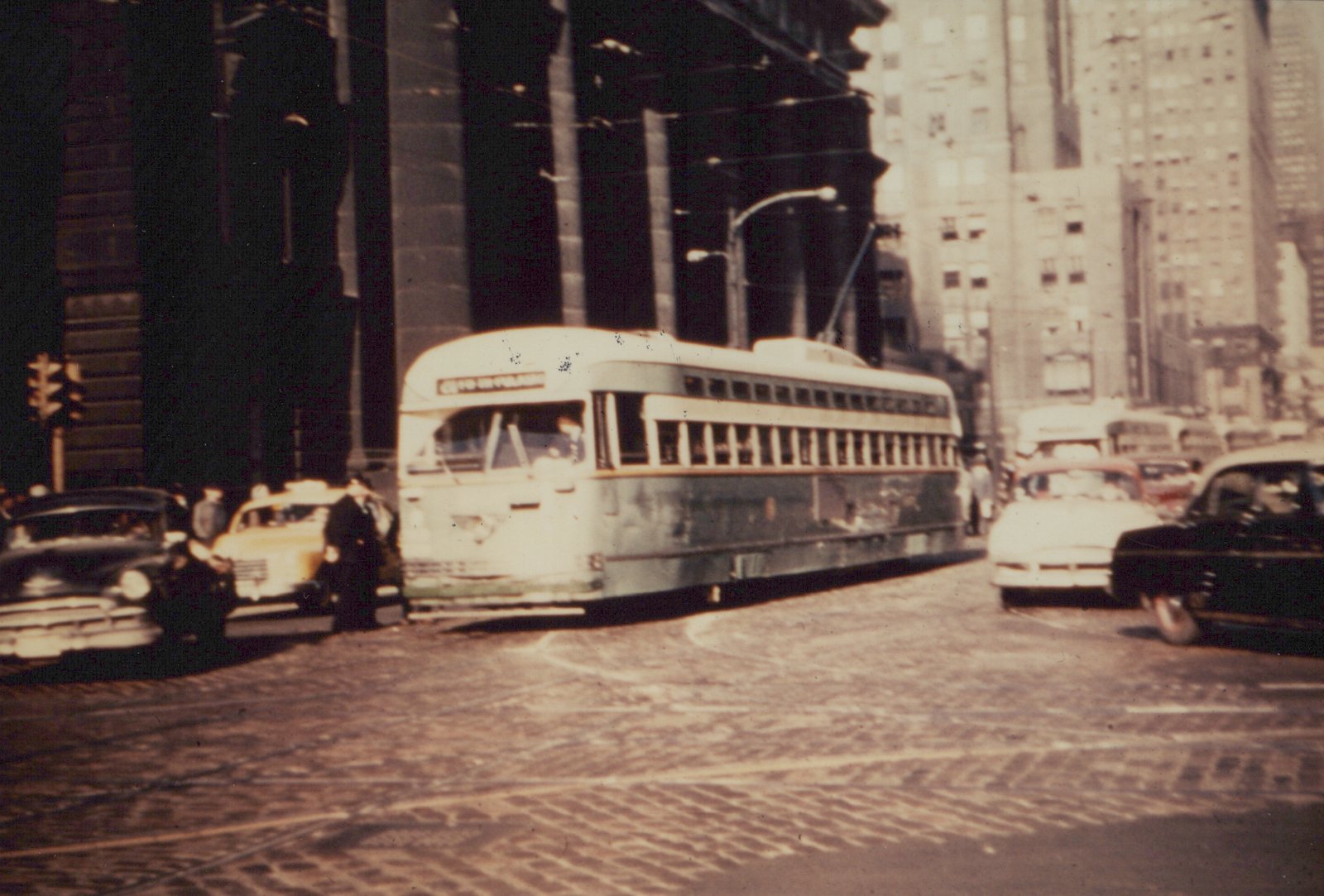 A green and cream streamlined streetcar with its trolley pole raised crosses a granite-paved intersection, with cars, a yellow taxi, a large columned building and high-rise buildings around it.