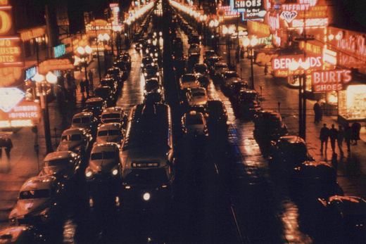 An elevated night view down a wet city street lined with neon store signs, with a bus in the center lane surrounded by taxicabs and 1950s automobiles and pedestrians on the sidewalks.