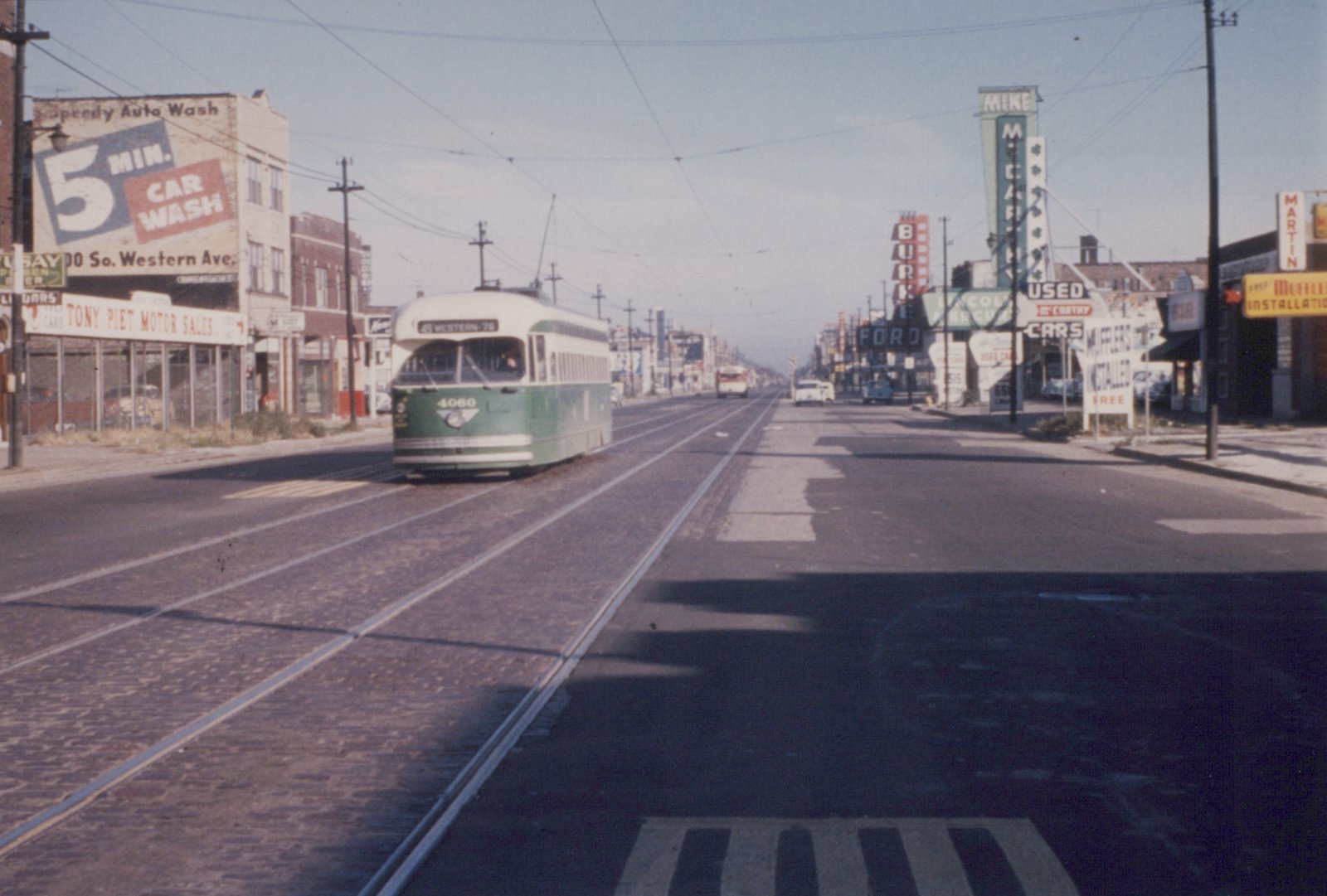 A green and cream streetcar comes toward the camera on brick-paved double track under overhead wire, on a wide street lined with used car lots and large signs.