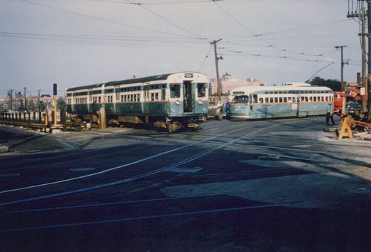 A green and cream two-car rapid transit train crossing a street with embedded streetcar tracks, next to a green and cream streetcar under overhead wires, with a man walking at right.