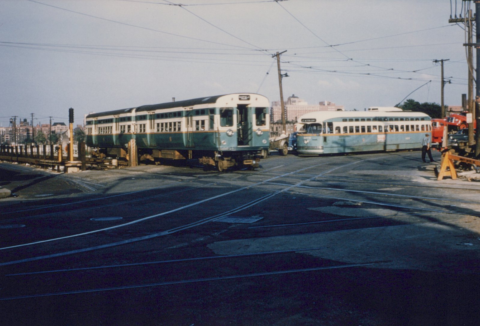 A green and cream two-car rapid transit train crossing a street with embedded streetcar tracks, next to a green and cream streetcar under overhead wires, with a man walking at right.