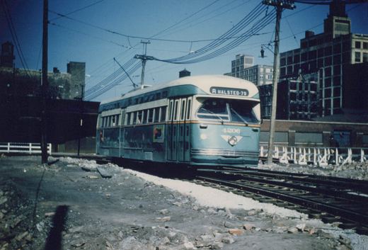 A green and cream streetcar numbered 4206, with a HALSTED-79 sign, travels on track laid over fresh ballast through a rubble-strewn lot, with overhead wires, wooden poles and brick industrial buildings behind it.