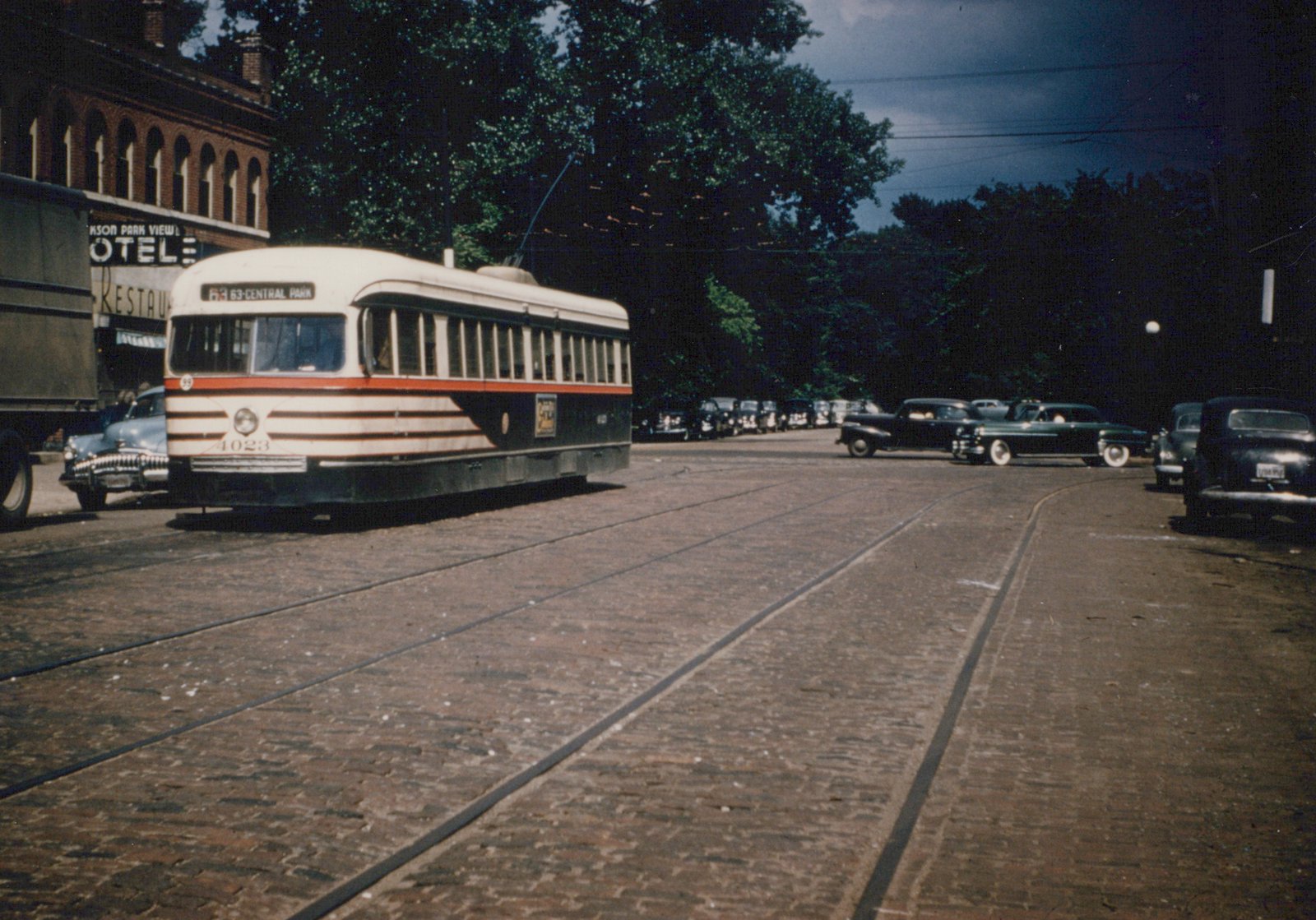 A green and cream streetcar with a red stripe, signed 63-CENTRAL PARK, rounds a curve on a brick street with streetcar tracks; a brick hotel building is at left, trees and parked 1940s cars are behind it, and a dark storm sky is overhead.