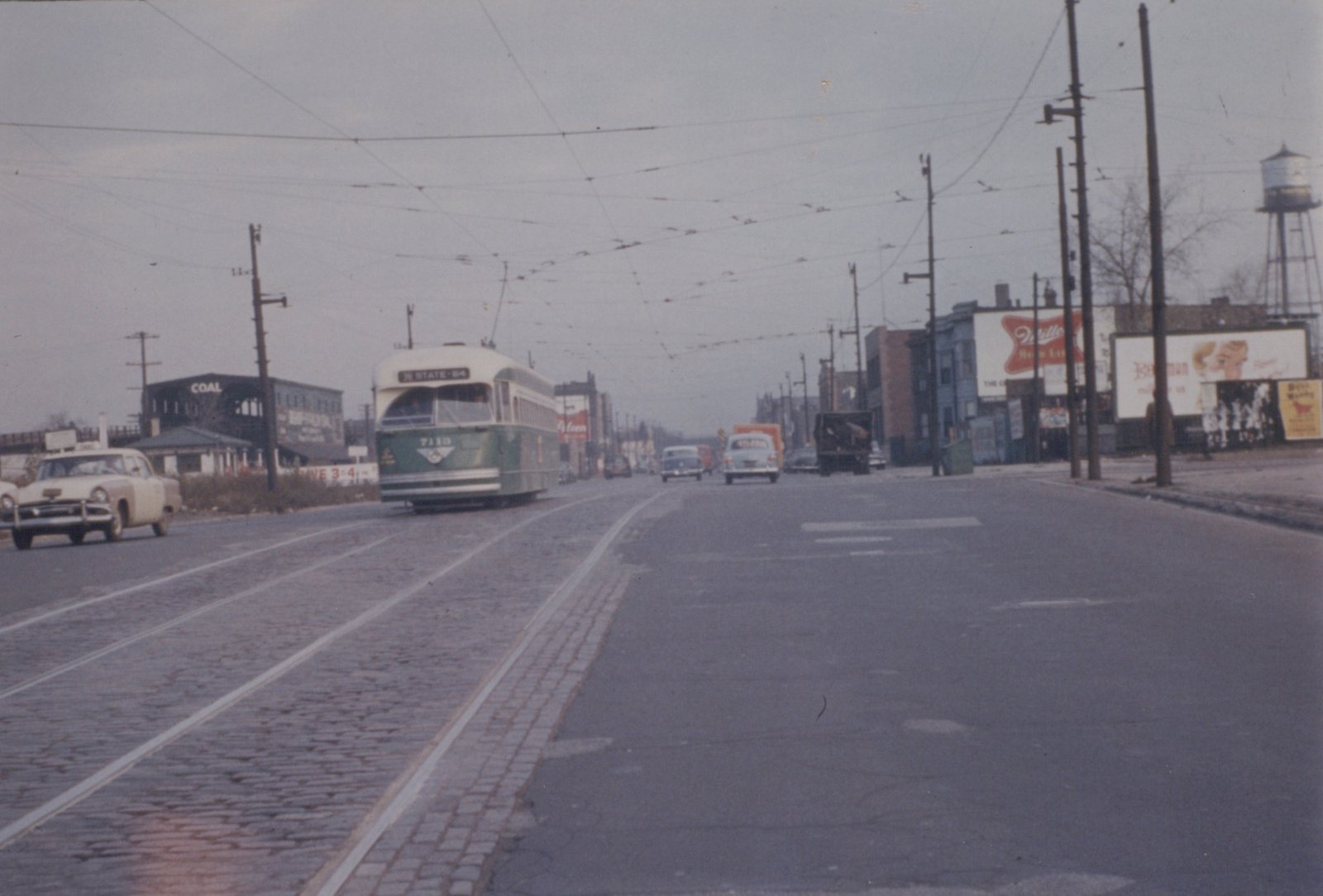 A green and cream streetcar drives toward the camera on tracks set in cobblestone paving, under a web of overhead trolley wires, with cars, billboards and a water tower along the street.