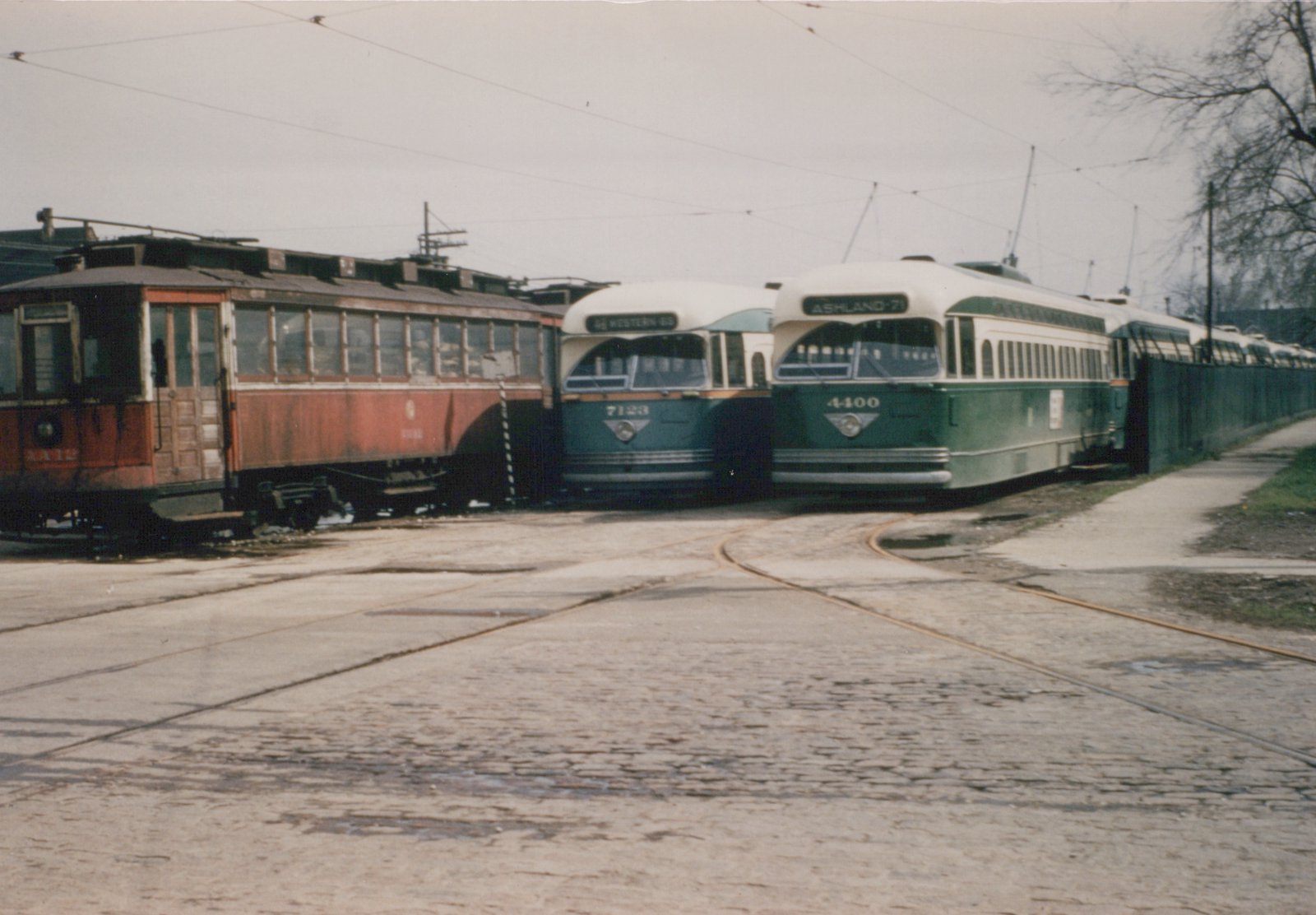 Two green and cream streamlined streetcars numbered 7123 and 4400 stand beside a faded red older streetcar on curved track in a cobblestone yard, with more streetcars lined up behind a green fence.
