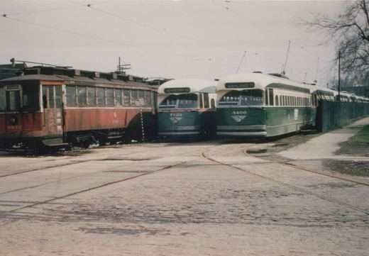 Two green and cream streamlined streetcars numbered 7123 and 4400 stand beside a faded red older streetcar on curved track in a cobblestone yard, with more streetcars lined up behind a green fence.