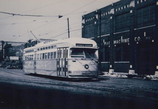 Black-and-white photo of a streamlined streetcar with its trolley pole raised, on a snowy street in front of a brick factory lettered MACHINE CO., with overhead wires above.