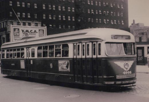 Black-and-white photo of a streamlined two-tone streetcar numbered 4008 with its trolley pole raised, moving past a tall brick building and a Y.M.C.A. billboard.