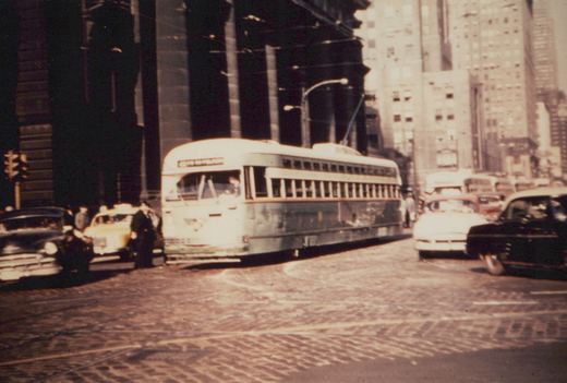 A green and cream streetcar with a trolley pole turns on a brick street next to a massive columned stone building. Cars of the early 1950s, including a yellow taxi, are in the street, with tall buildings behind.