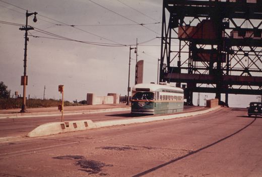 A green and cream streetcar with a single trolley pole runs on a wide, curving bridge approach beneath a massive dark steel truss frame. A traffic signal stands on a concrete median, and a dark old sedan drives away at the right.