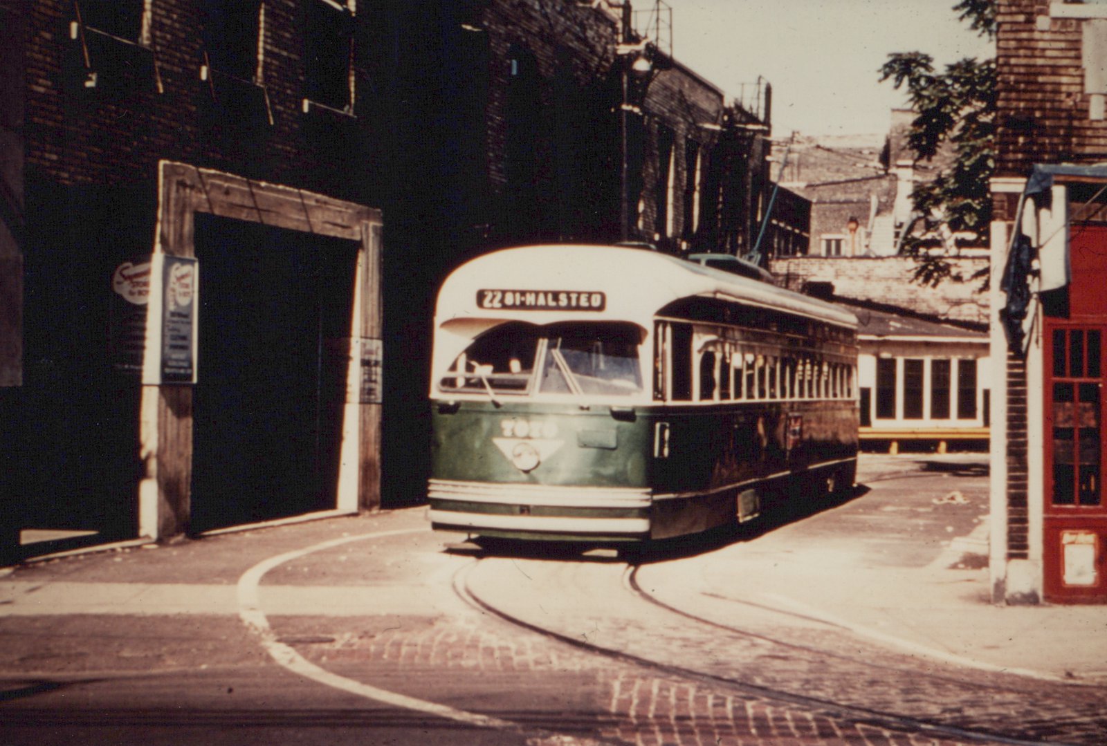 A green and cream streetcar with a rounded front and a trolley pole turns on a curved brick-paved track between dark brick buildings on a sunny day.