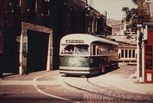 A green and cream streetcar with a rounded front and a trolley pole turns on a curved brick-paved track between dark brick buildings on a sunny day.