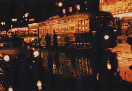 A green and cream streamlined streetcar on a wet street at night, with its headlight on and a roll sign reading 36 Broadway Devon. Two people stand beside it, and a lit theater marquee advertising Judy Holliday is at upper right.