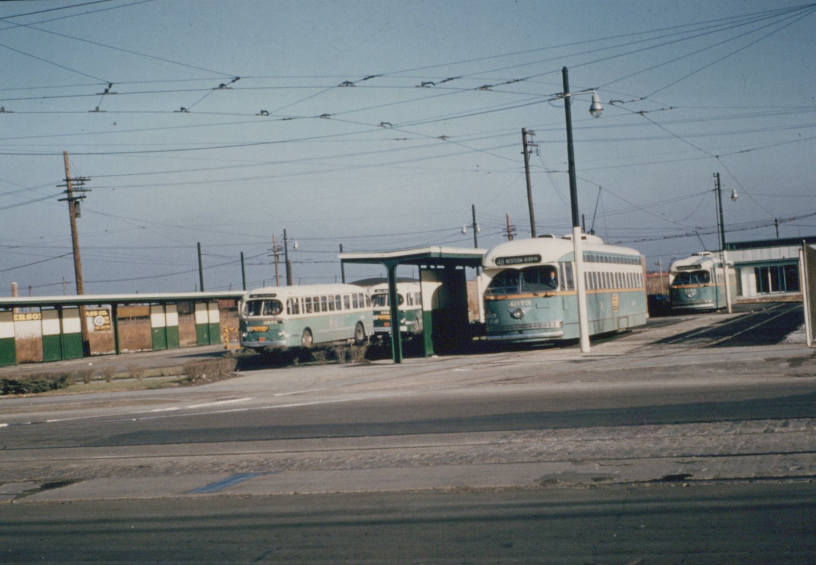 A green and cream streetcar with a lit 49 Western-Berwyn sign stands under a web of overhead wires beside a passenger shelter, with another streetcar to the right and two buses to the left.