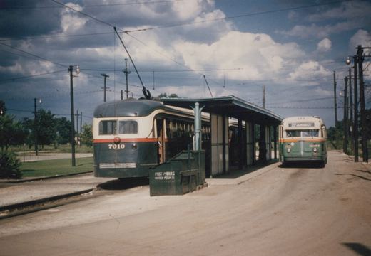A green and cream streetcar with its trolley pole raised stands beside a covered shelter, and a green and cream bus is parked on a dirt lane on the shelter's other side, under a cloudy blue sky.