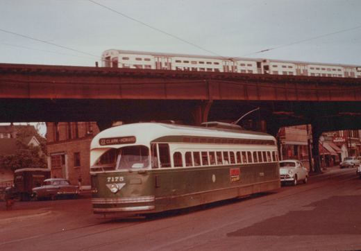 A green and cream streetcar signed 22 Clark-Howard runs along a street under a rust-colored steel elevated structure, with a train of light-colored double-deck commuter coaches on top and parked cars and storefronts beyond.