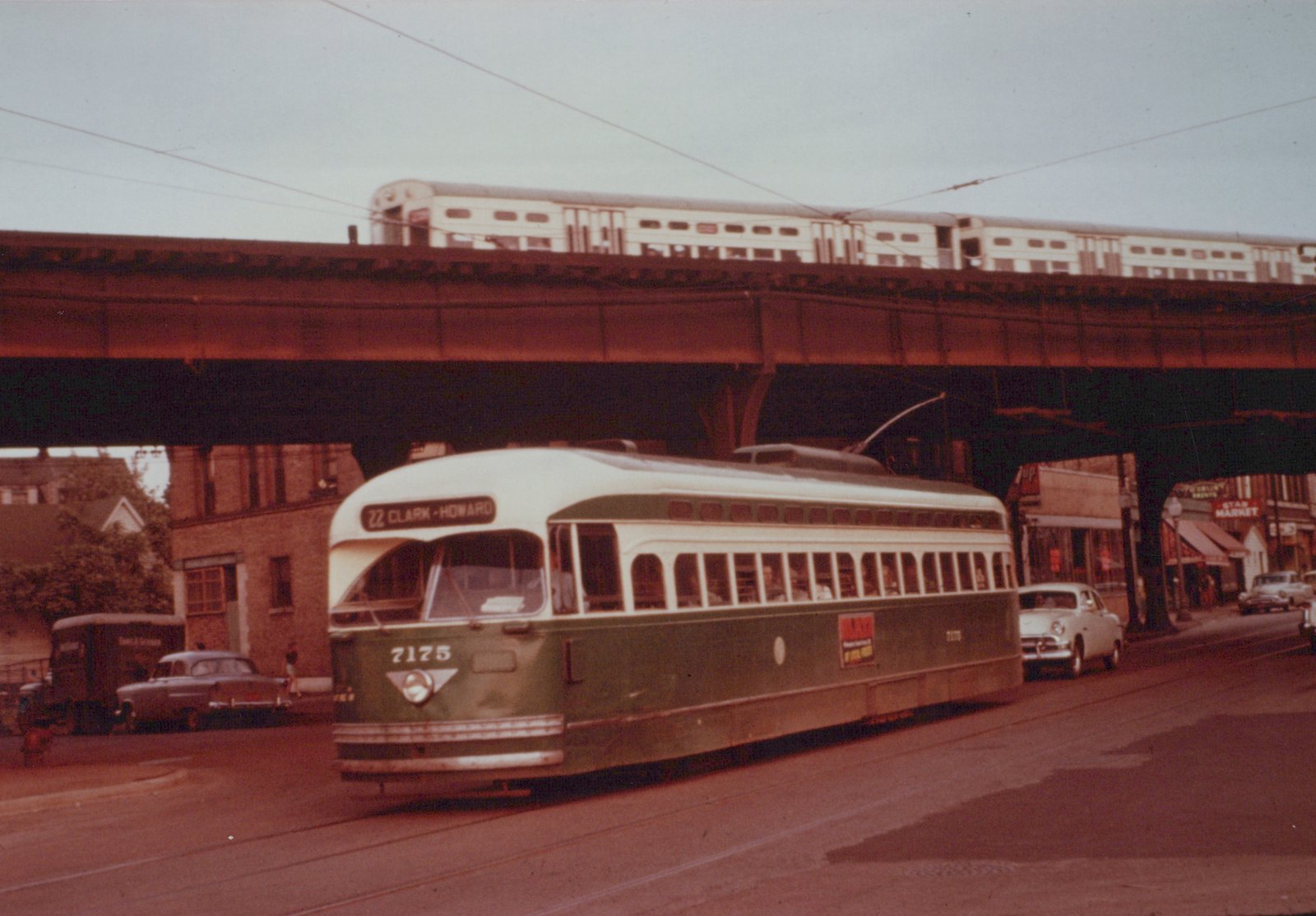 A green and cream streetcar signed 22 Clark-Howard runs along a street under a rust-colored steel elevated structure, with a train of light-colored double-deck commuter coaches on top and parked cars and storefronts beyond.