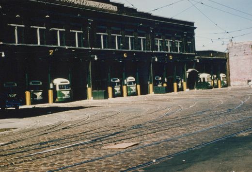 A dark brick two-story carbarn with numbered open bays holding about ten green and cream streamlined streetcars, with curving tracks set in brick pavement and overhead wires in the foreground.