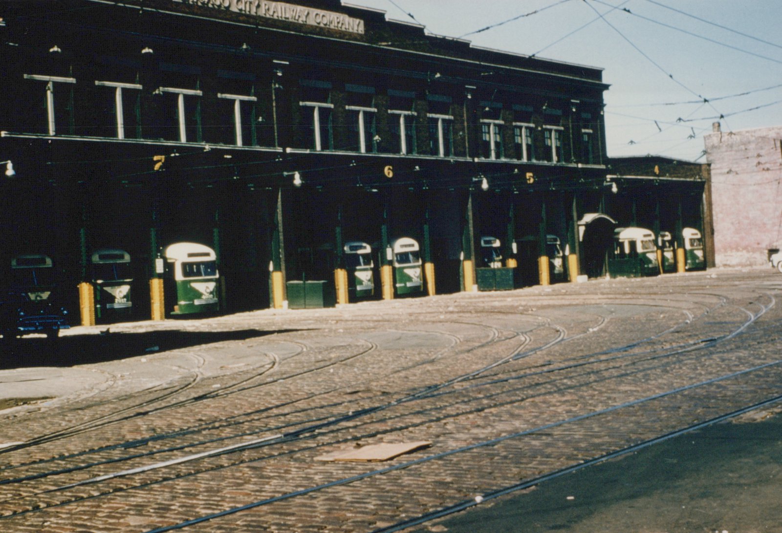 A dark brick two-story carbarn with numbered open bays holding about ten green and cream streamlined streetcars, with curving tracks set in brick pavement and overhead wires in the foreground.