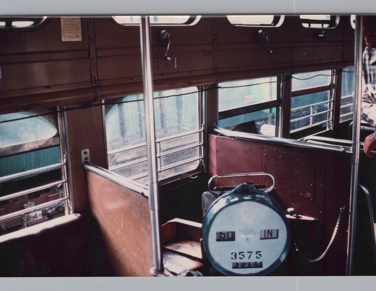 Inside a streetcar, a round farebox numbered 3575 sits in a railed conductor's area beside brown seat backs. Barred windows sit below a row of small upper windows, and a man stands holding a railing at the right edge.