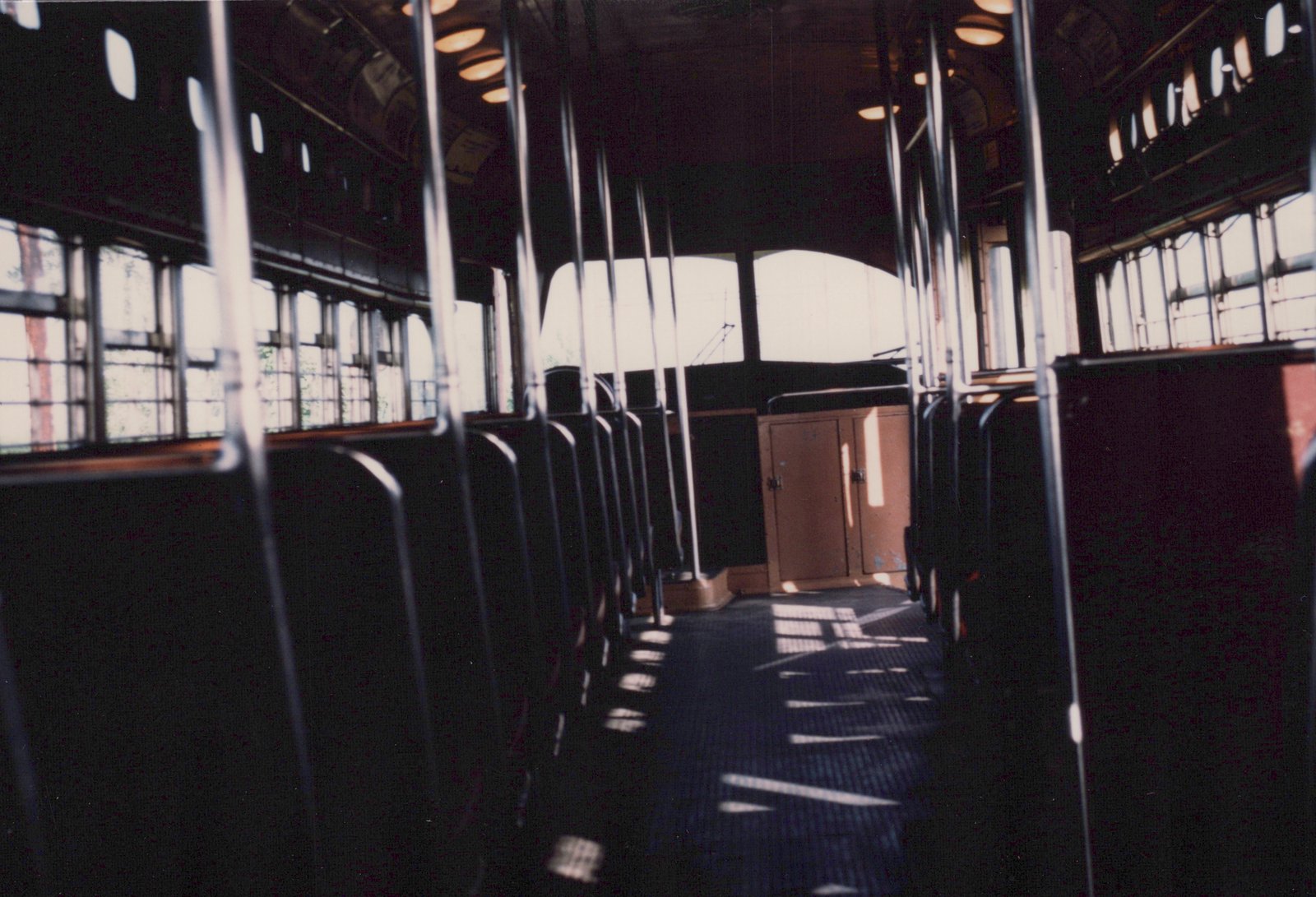 Dim streetcar interior with rows of dark seats and chrome grab handles along a sunlit aisle. At the far end is a cab with an orange cabinet under two bright end windows, and a trolley pole shows outside.