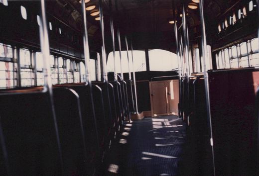 Dim streetcar interior with rows of dark seats and chrome grab handles along a sunlit aisle. At the far end is a cab with an orange cabinet under two bright end windows, and a trolley pole shows outside.