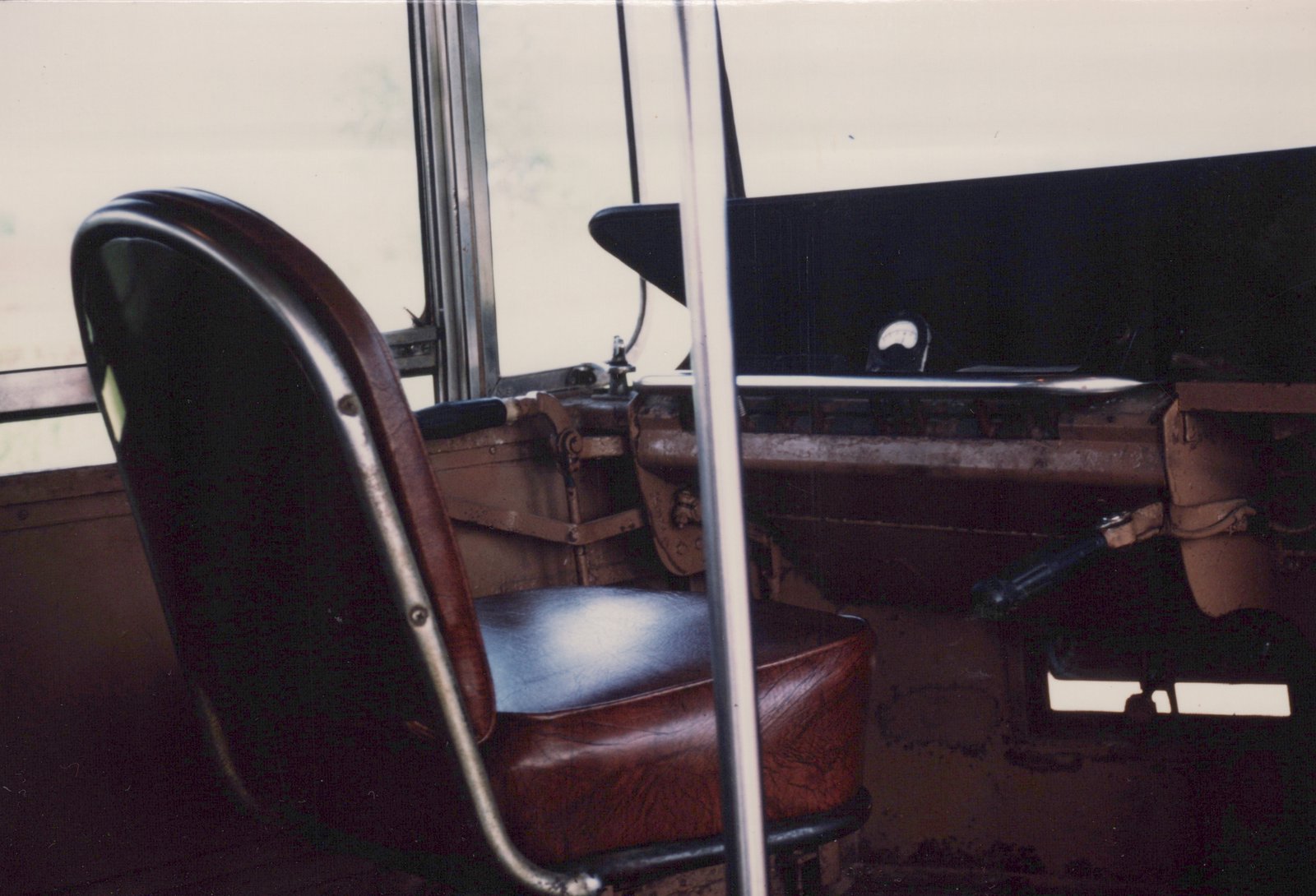 Close view from inside a streetcar of a brown leather operator's seat on a chrome frame beside brown-painted control gear, a black dash panel with a small dial meter, and bright windows behind.