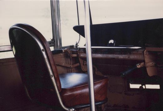 Close view from inside a streetcar of a brown leather operator's seat on a chrome frame beside brown-painted control gear, a black dash panel with a small dial meter, and bright windows behind.