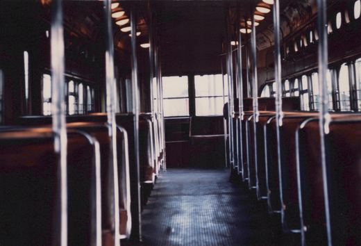 A dim, empty interior of an old railway car: a center aisle with a ribbed floor runs between rows of brown seats, each with a vertical metal grab pole, under a row of ceiling lights, with arched windows along the sides and a pair of large windows at the far end.