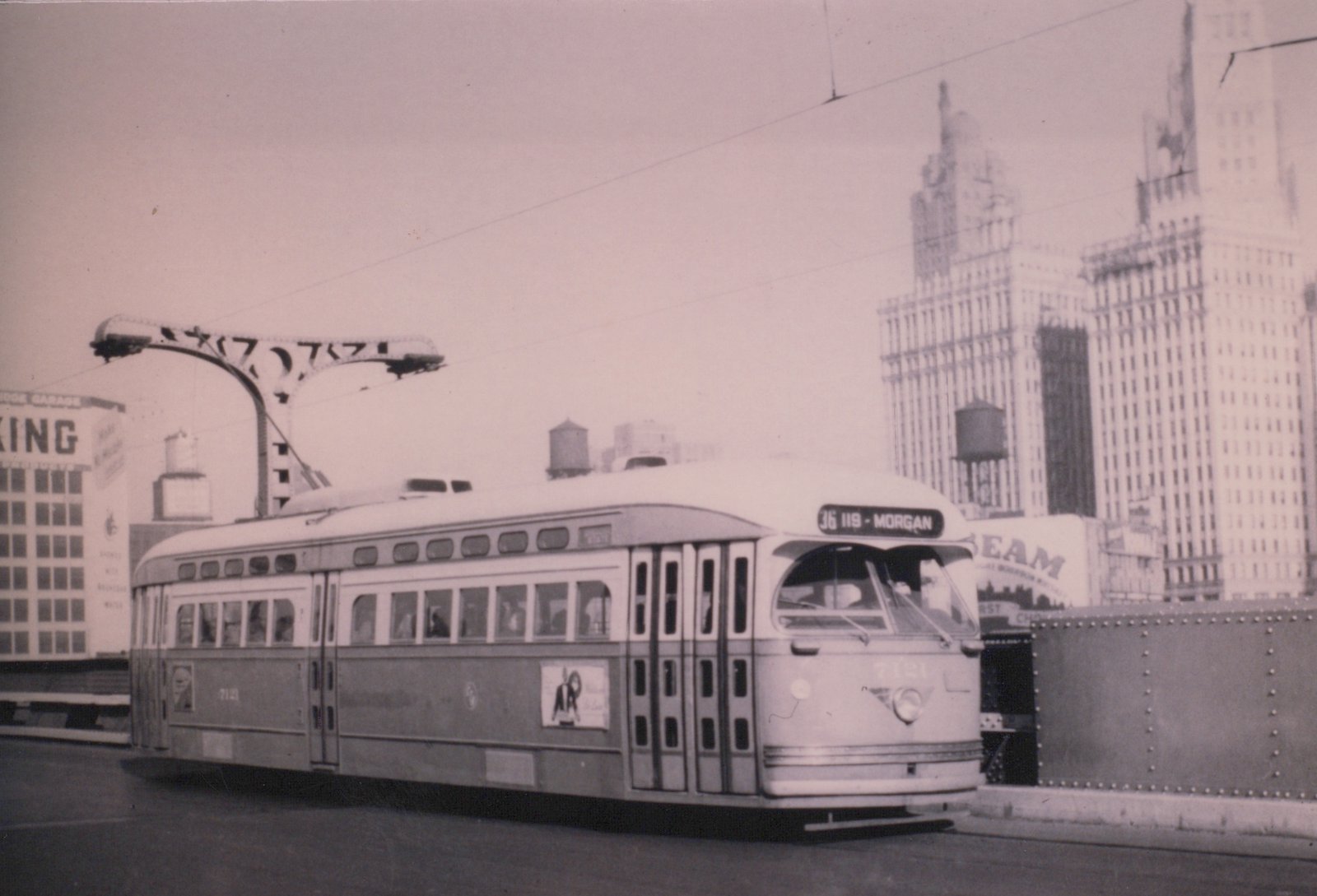 Black-and-white photo of a two-tone streamlined streetcar signed 36 119 - Morgan on a bridge under overhead wires, with a riveted steel railing, an ornate street light and tall downtown office towers behind it.