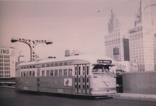 Black-and-white photo of a two-tone streamlined streetcar signed 36 119 - Morgan on a bridge under overhead wires, with a riveted steel railing, an ornate street light and tall downtown office towers behind it.