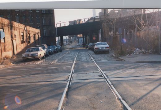 A cobblestone street lined with parked 1980s cars and brick industrial buildings. Two rail tracks run down its center and cross other tracks in the paved foreground, and a dark steel overpass spans the street.