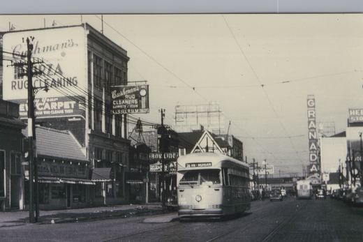 Black-and-white street scene with a streamlined streetcar under overhead wires, large rug cleaner signs on buildings at left and a tall GRANADA sign down the street.