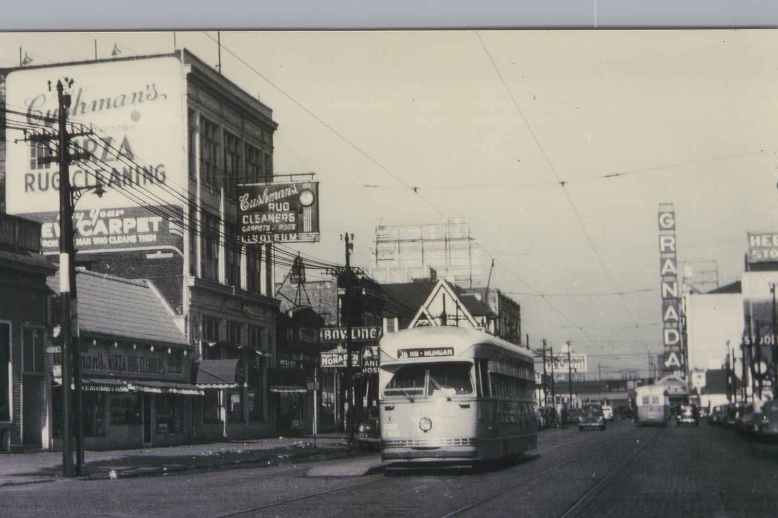 Black-and-white street scene with a streamlined streetcar under overhead wires, large rug cleaner signs on buildings at left and a tall GRANADA sign down the street.