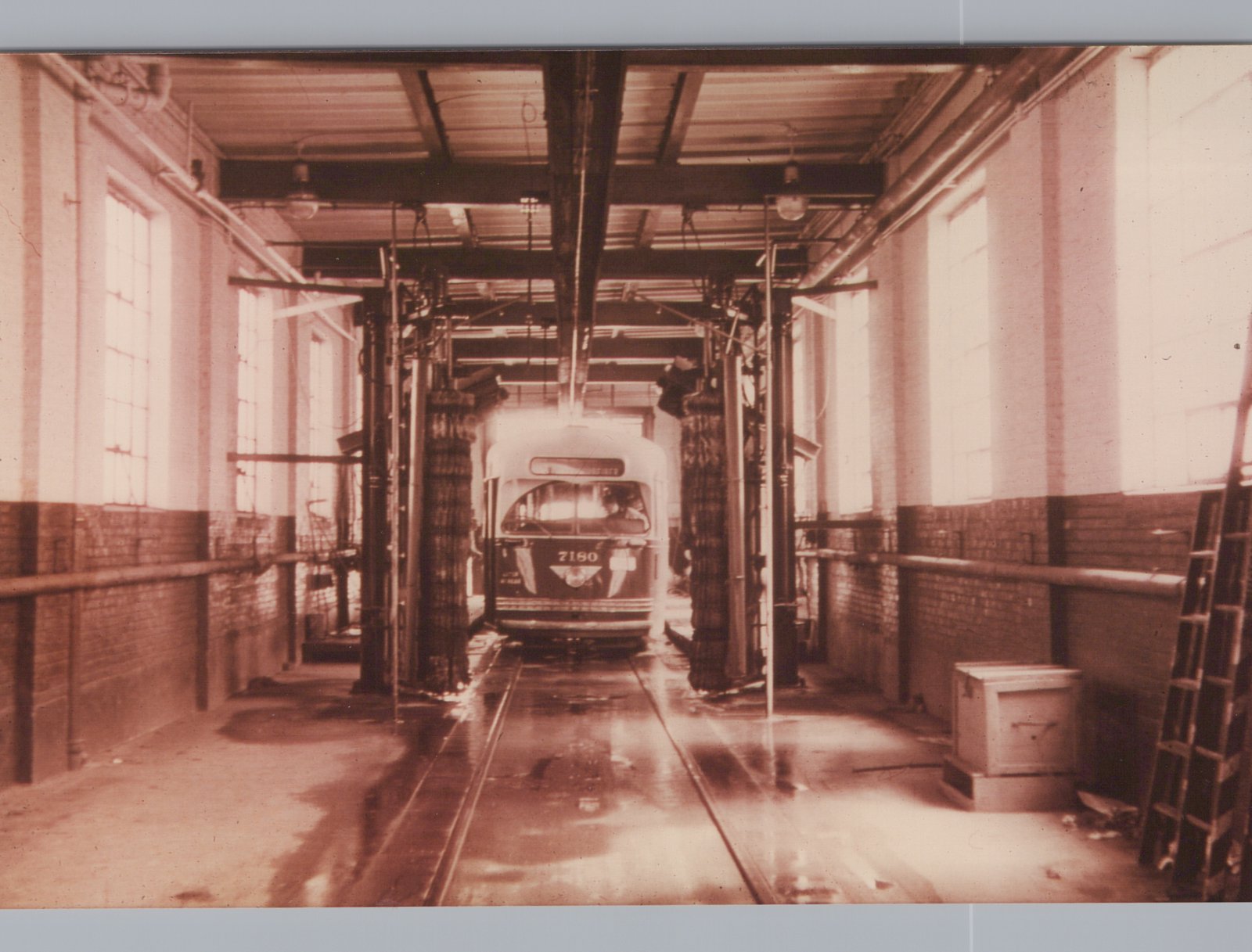 Sepia photo of a streetcar numbered 7180 entering an indoor car wash, between tall rotating brushes on a wet floor with track, under a steel-beamed roof with brick walls and tall windows.