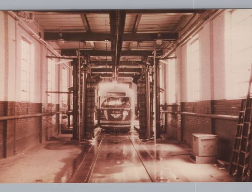 Sepia photo of a streetcar numbered 7180 entering an indoor car wash, between tall rotating brushes on a wet floor with track, under a steel-beamed roof with brick walls and tall windows.