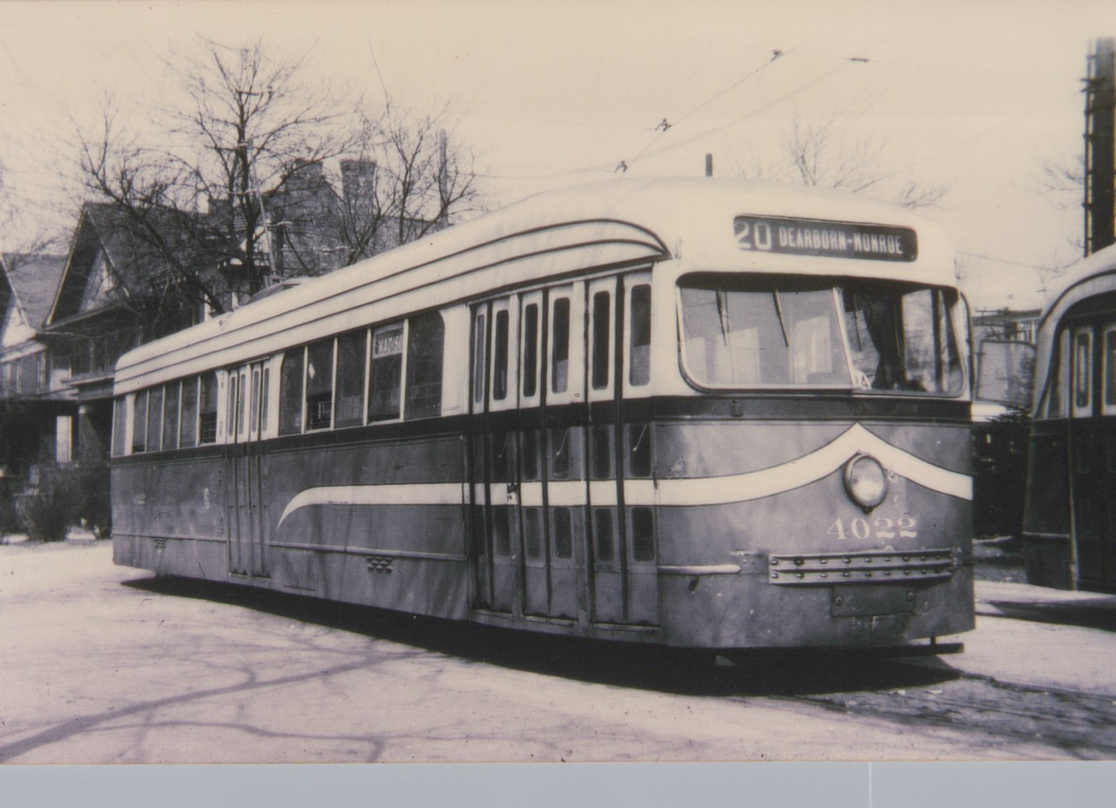 A black and white photo of a streamlined streetcar with a dark lower body and white upper body. It has a cream stripe sweeping up over its single headlight, the number 4022 on the front, and a sign reading 20 Dearborn-Monroe. It stands on pavement in front of bare trees and houses, and a second stre