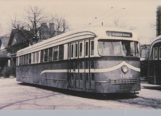 A black and white photo of a streamlined streetcar with a dark lower body and white upper body. It has a cream stripe sweeping up over its single headlight, the number 4022 on the front, and a sign reading 20 Dearborn-Monroe. It stands on pavement in front of bare trees and houses, and a second stre