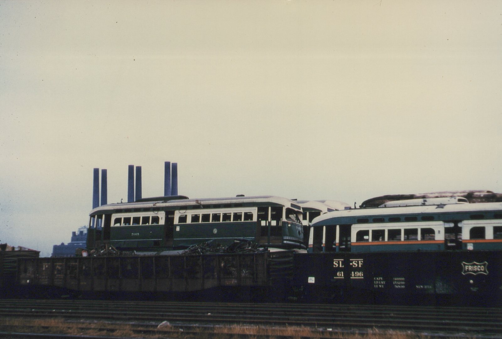 Two stripped green-and-cream streetcars with their windows removed sit among scrap metal behind a black Frisco gondola and rail tracks, with six smokestacks against an overcast sky.