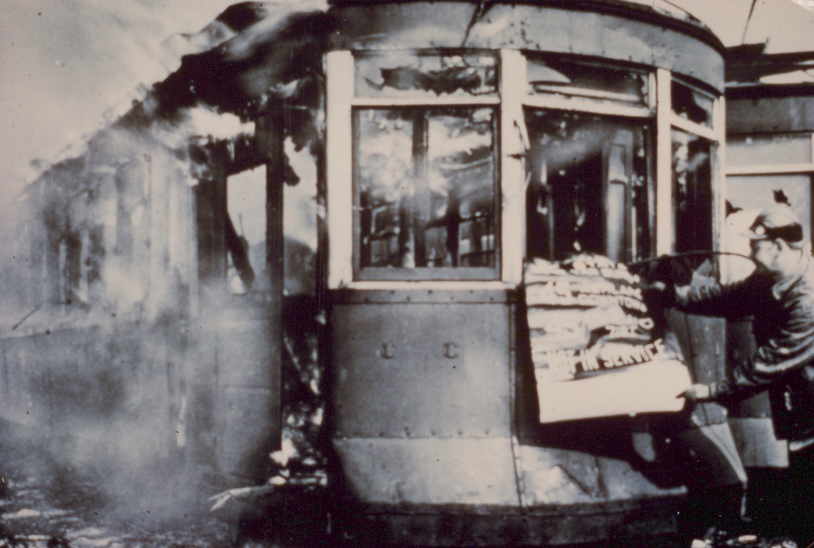 Black and white photo of a damaged streetcar with a curved front and large windows, with smoke rising from its side. A person in uniform holds up a sign that says 'NOT IN SERVICE'.