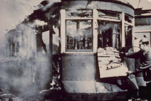 Black and white photo of a damaged streetcar with a curved front and large windows, with smoke rising from its side. A person in uniform holds up a sign that says 'NOT IN SERVICE'.