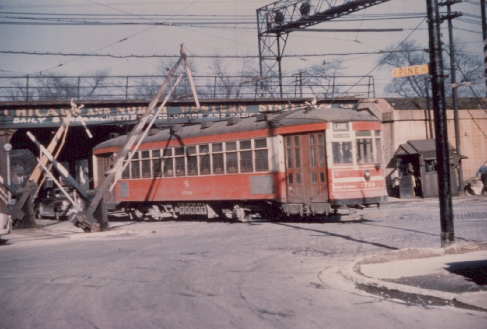 A red and cream interurban car, number 1766, is stopped at a railroad crossing under a large bridge with the words 'Chicago and North Western System' on it; the street sign reads 'Pine Av' and the car's destination sign reads 'LAKE'.