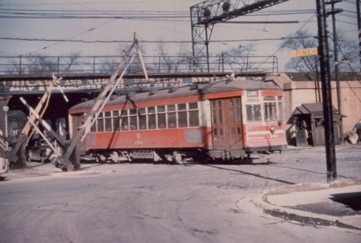 A red and cream interurban car, number 1766, is stopped at a railroad crossing under a large bridge with the words 'Chicago and North Western System' on it; the street sign reads 'Pine Av' and the car's destination sign reads 'LAKE'.