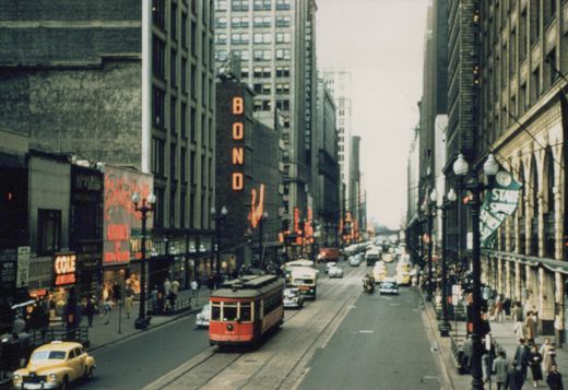 A red Birney streetcar marked 351 travels down a busy State Street in Chicago, flanked by buildings with signs for 'BOND' and 'COLE JEWELERS', with vintage cars and pedestrians on the sidewalks.