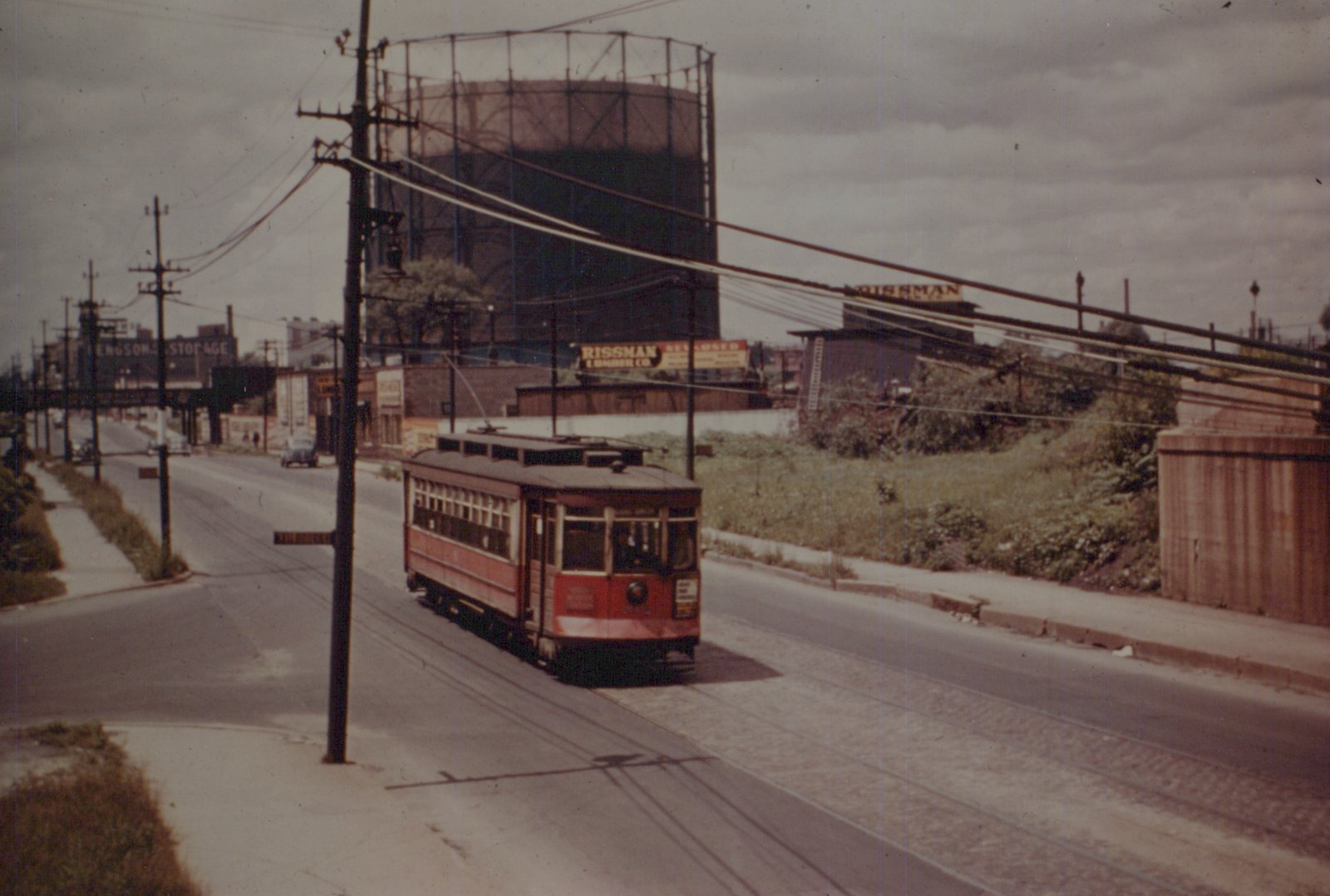 A red and cream wooden streetcar runs down a city street under overhead wires, with a large gas holder and industrial buildings in the background.